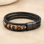 Gros plan d'un bracelet homme en cuir tressé noir avec perles d'œil de tigre sur fond pierre claire