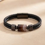 Gros plan d'un bracelet homme en cuir tressé noir avec perle d'agate marron sur fond pierre claire