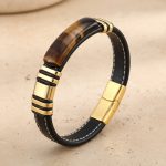 Gros plan d'un bracelet homme en cuir plat noir surpiqué avec plaque en œil de tigre et acier doré sur fond pierre claire