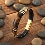 Bracelet homme en cuir plat noir et plaque d'œil de tigre avec acier doré présenté sur bois