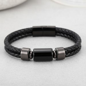 Gros plan d'un bracelet homme en cuir tressé noir avec perle cylindrique en acier noir sur fond pierre claire
