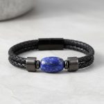 Gros plan d'un bracelet homme en cuir tressé noir avec perle de lapis-lazuli sur fond pierre claire