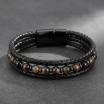 Gros plan d'un bracelet homme en cuir tressé noir avec perles d'œil de tigre foncé sur fond pierre sombre