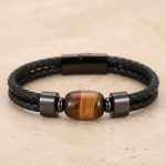 Gros plan d'un bracelet homme en cuir tressé noir avec perle d'œil de tigre sur fond pierre claire
