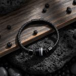 Bracelet homme en cuir tressé noir et pierre de lave présenté sur roche volcanique et bois