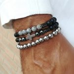 Poignet d'un homme portant un ensemble de trois bracelets élastiques en perles noires, hématite argentée et croix