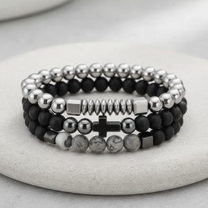 Gros plan d'un ensemble de trois bracelets homme en perles noires, hématite argentée et jaspe paysage gris sur fond pierre claire