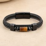 Gros plan d'un bracelet homme en cuir noir tressé et surpiqué avec tube en œil de tigre sur fond pierre claire