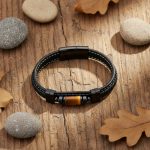 Bracelet homme en cuir noir et tube d'œil de tigre présenté sur bois avec éléments naturels