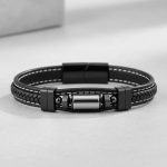 Gros plan d'un bracelet homme en cuir noir tressé et surpiqué avec tube en acier argenté sur fond pierre claire