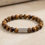 Gros plan d'un bracelet homme en perles d'œil de tigre doré avec barre en acier brossé sur fond pierre claire