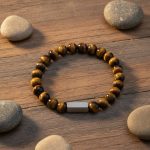 Bracelet homme en œil de tigre doré et barre en acier brossé présenté sur bois avec éléments naturels