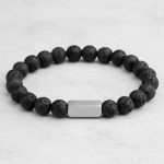 Gros plan d'un bracelet homme en perles de pierre de lave noire avec barre en acier brossé sur fond pierre claire
