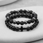 Gros plan d'un ensemble de deux bracelets homme en onyx noir, pierre de lave, hématite et croix sur fond pierre claire
