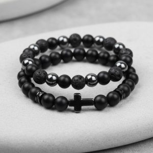 Gros plan d'un ensemble de deux bracelets homme en onyx noir, pierre de lave, hématite et croix sur fond pierre claire