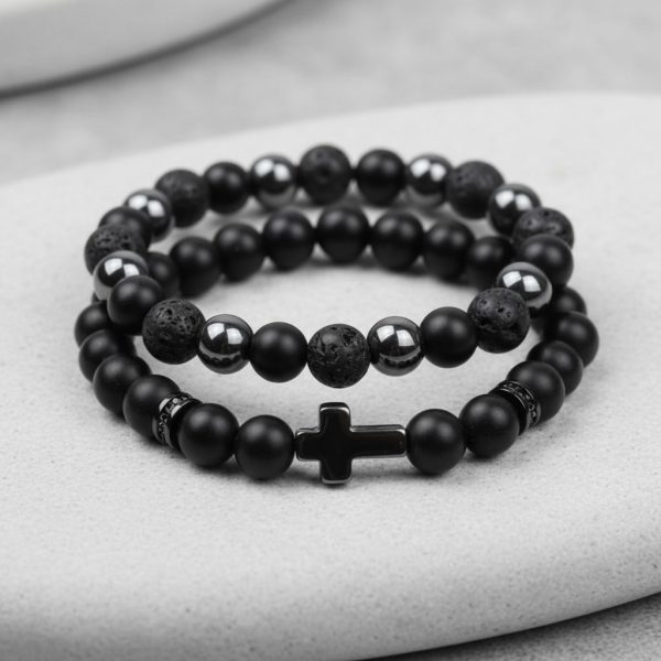 Gros plan d'un ensemble de deux bracelets homme en onyx noir, pierre de lave, hématite et croix sur fond pierre claire