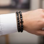 Poignet d'un homme portant un ensemble de deux bracelets élastiques en onyx noir, pierre de lave, hématite et croix