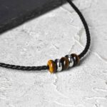 ollier cuir noir homme perles œil de tigre marron rondelles acier argenté cordon tressé gros plan fond pierre