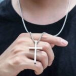 Collier pendentif croix minimaliste homme acier inoxydable argenté tenu en main t-shirt noir