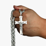 Main tenant un collier pendentif crucifix en acier inoxydable argenté avec chaîne maille byzantine fond blanc