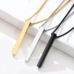 Collier pendentif homme barre acier inoxydable trois coloris doré argenté et noir posés à plat sur surface blanche